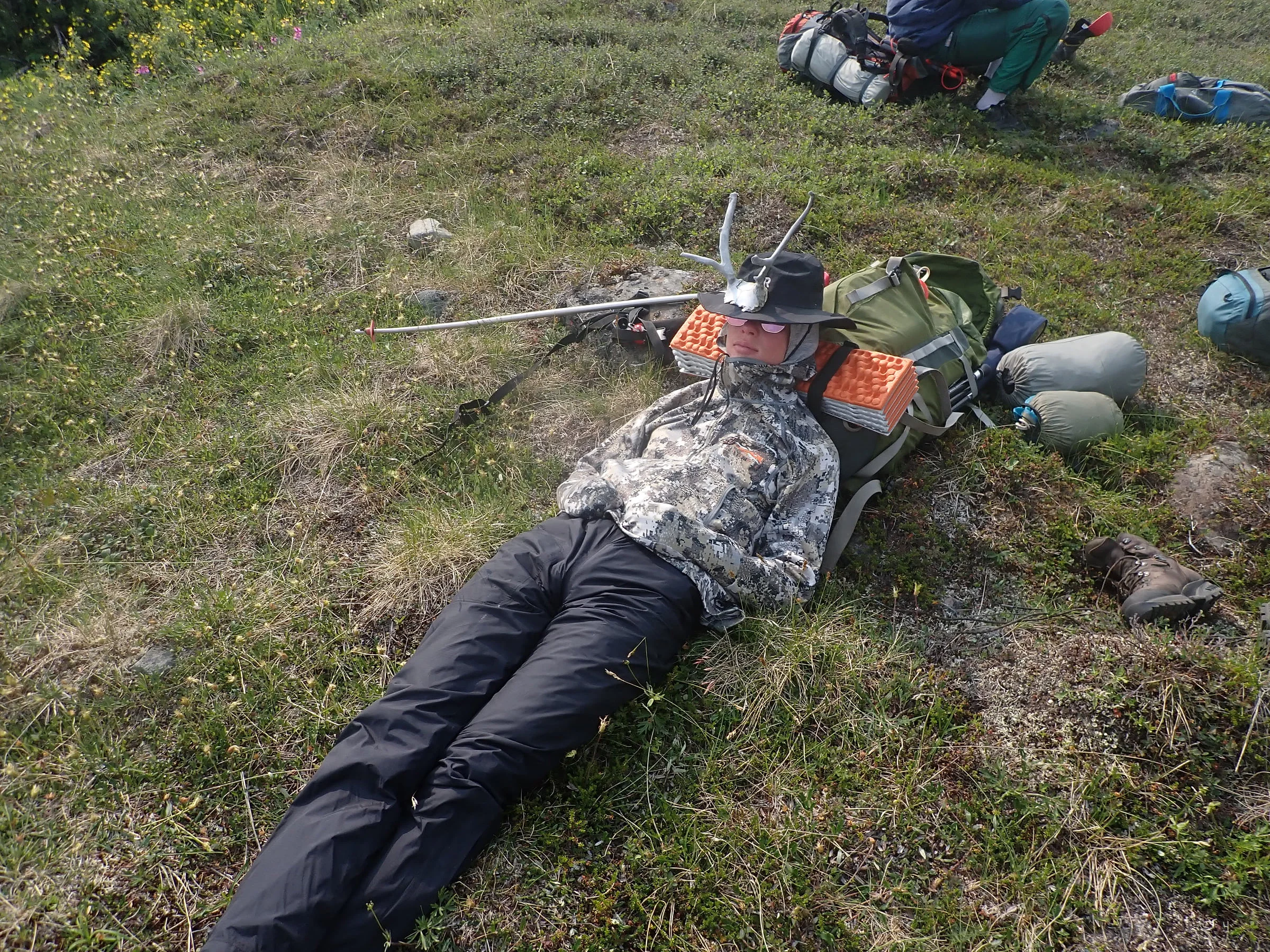 A backpacker wearing antlers on a wide-brimmed hat naps against a pack in the tundra.