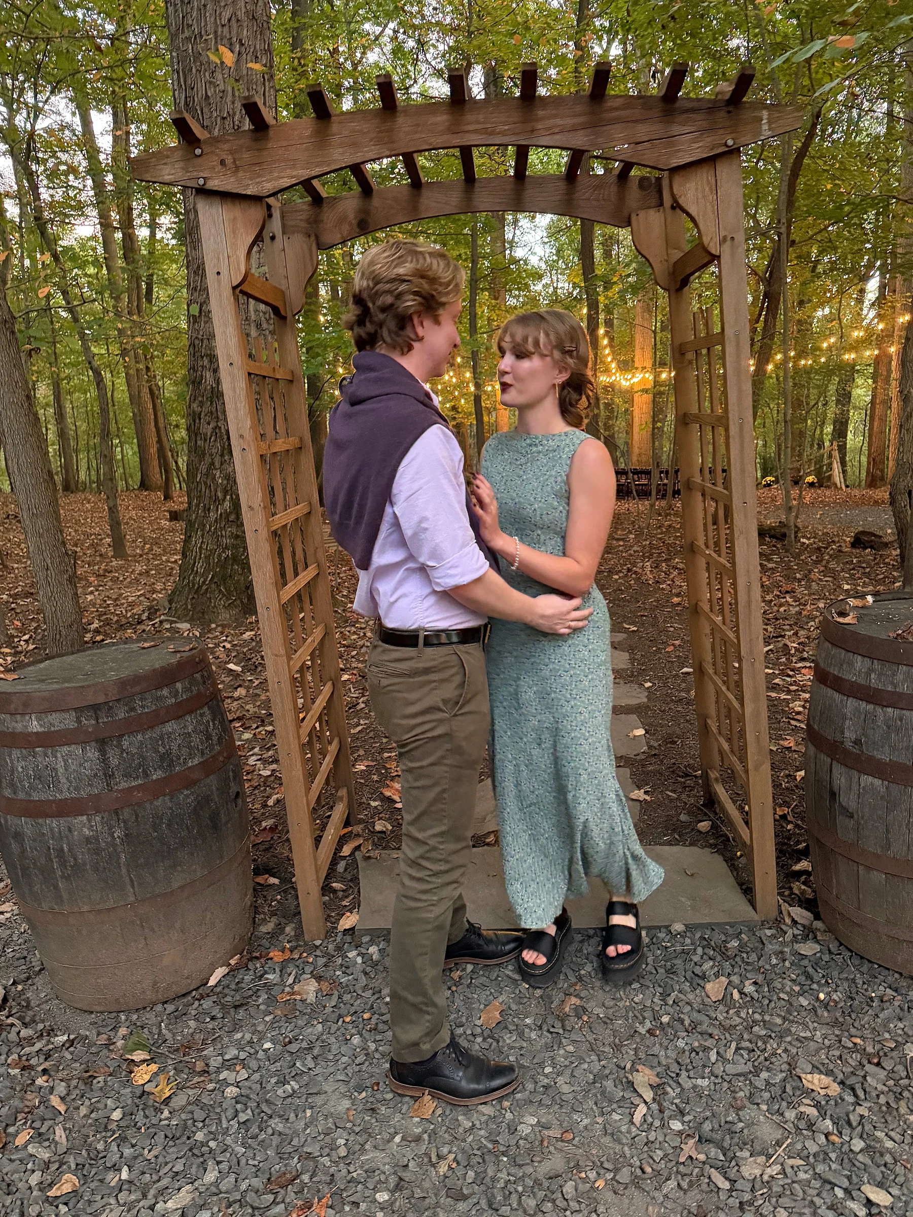 A formally dressed couple embraces beneath a wooden arbor strung with warm lights.