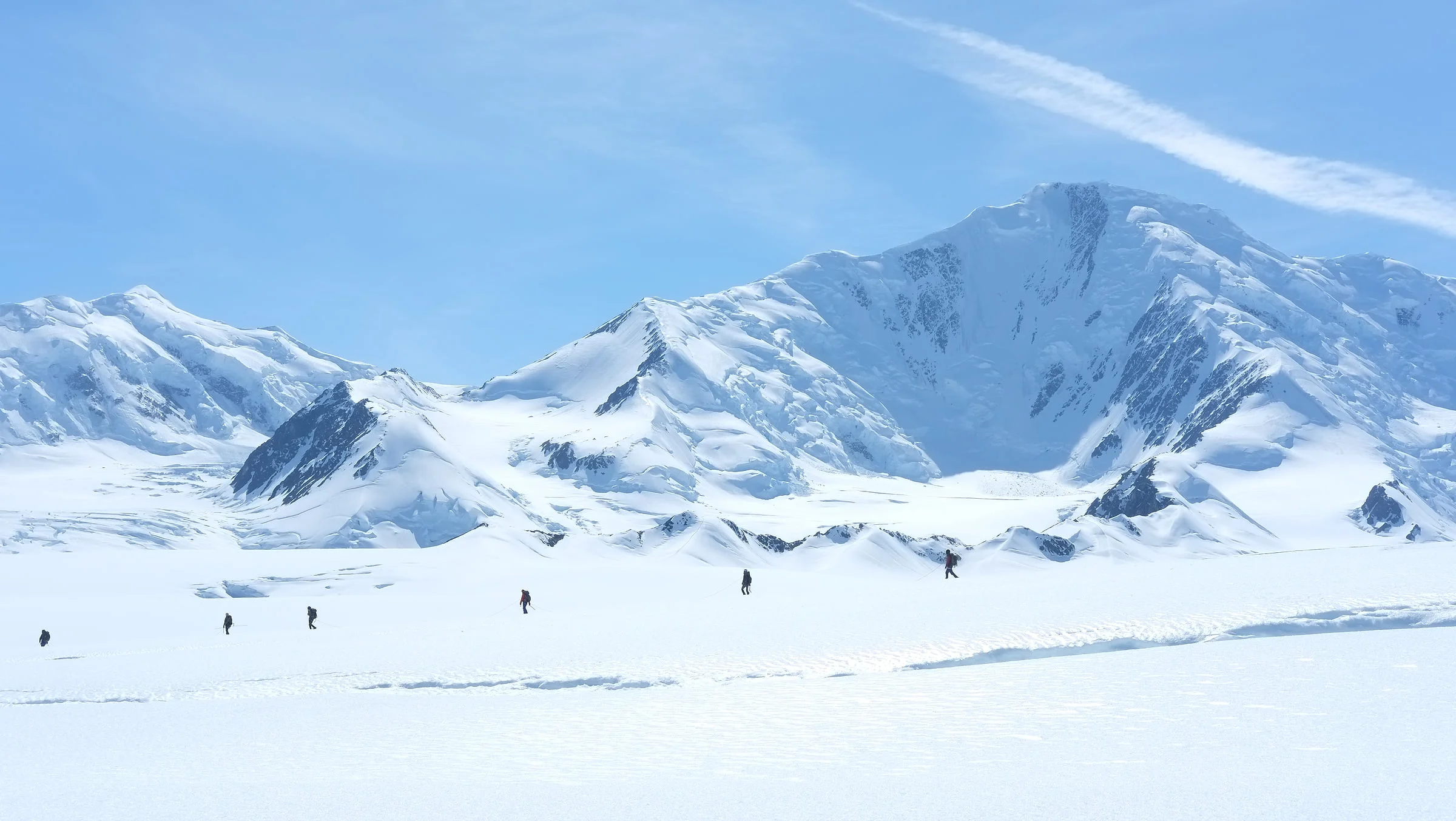 Tiny mountaineers cross a bright snowfield below immense glacier-covered peaks.