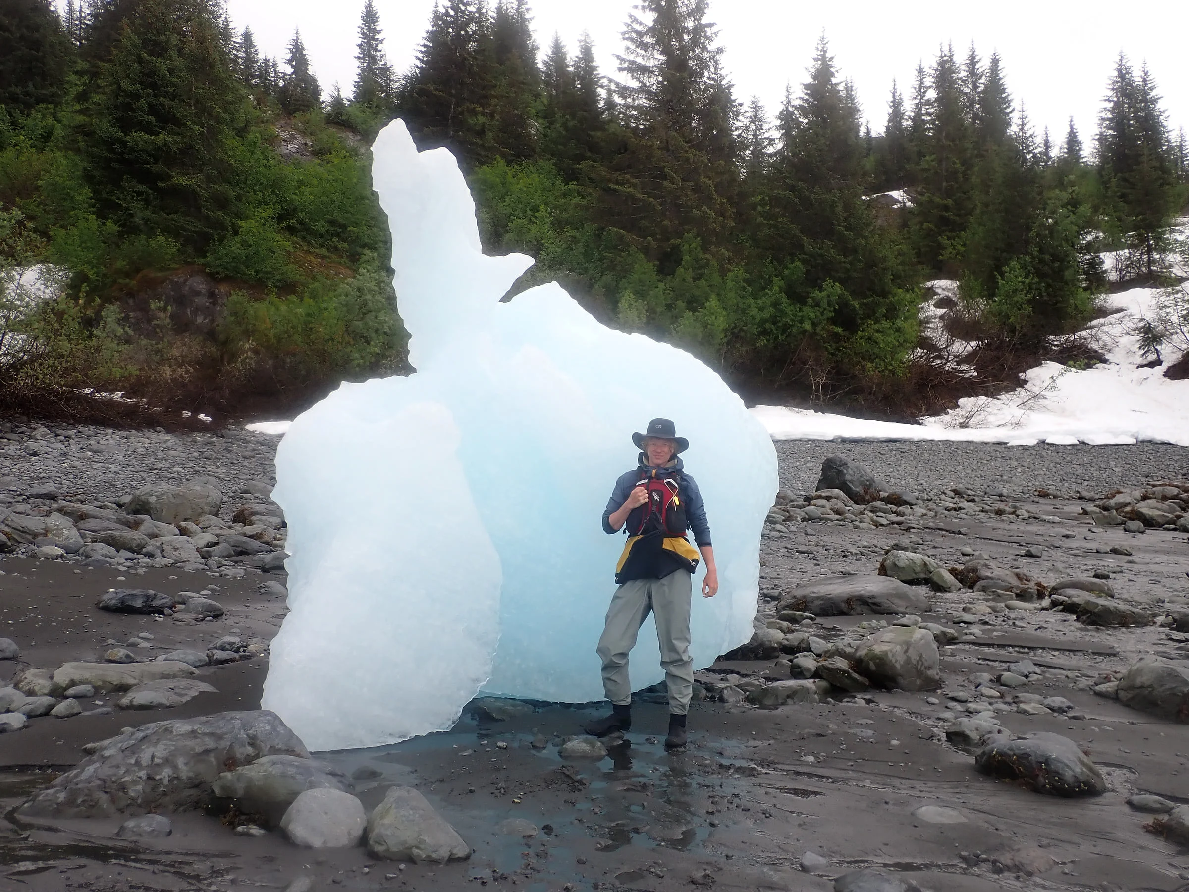 A hiker stands beside a towering blue-white chunk of ice on a rocky shore.