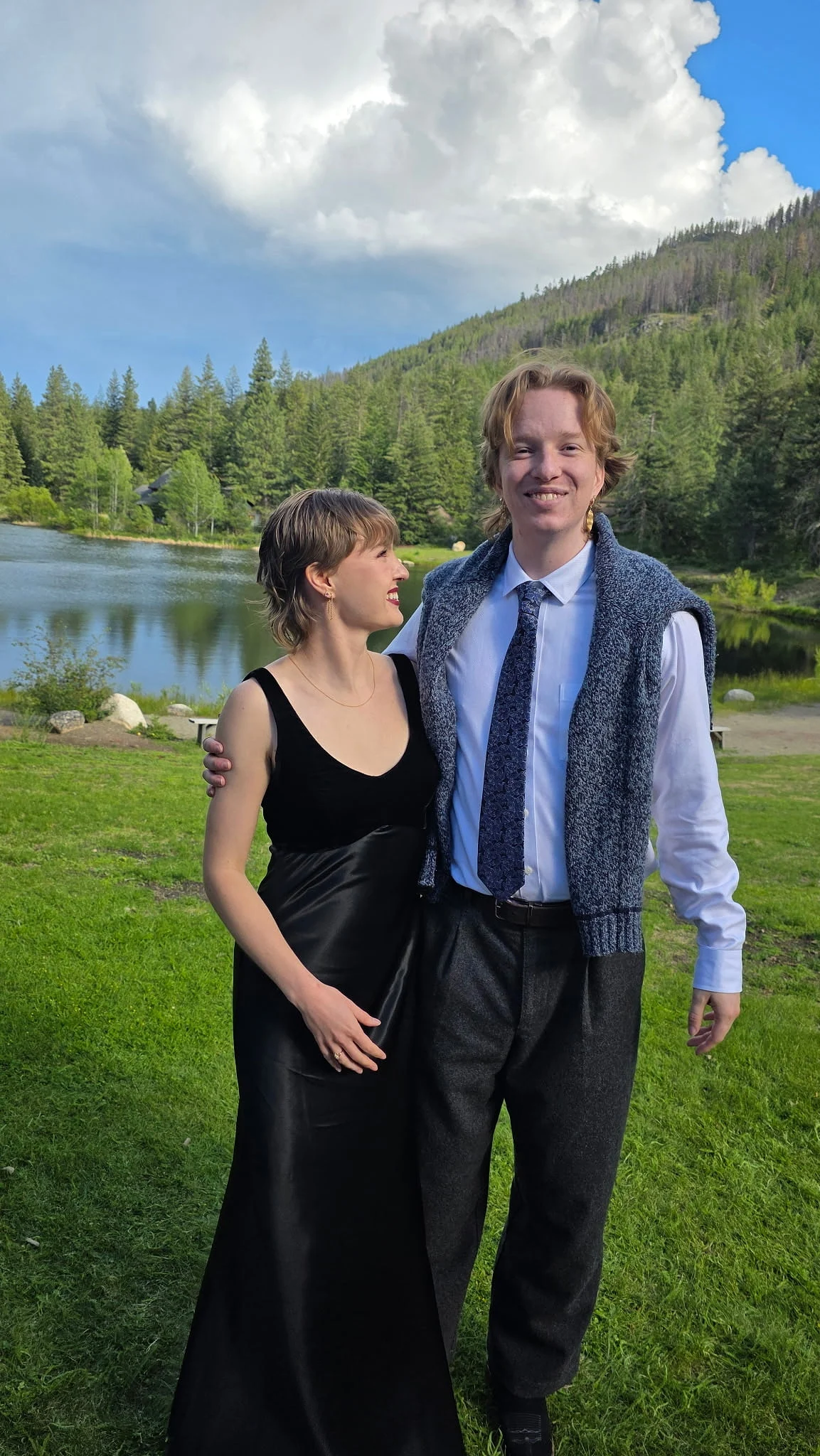 A couple dressed for an event stands arm in arm beside a mountain lake beneath towering clouds.