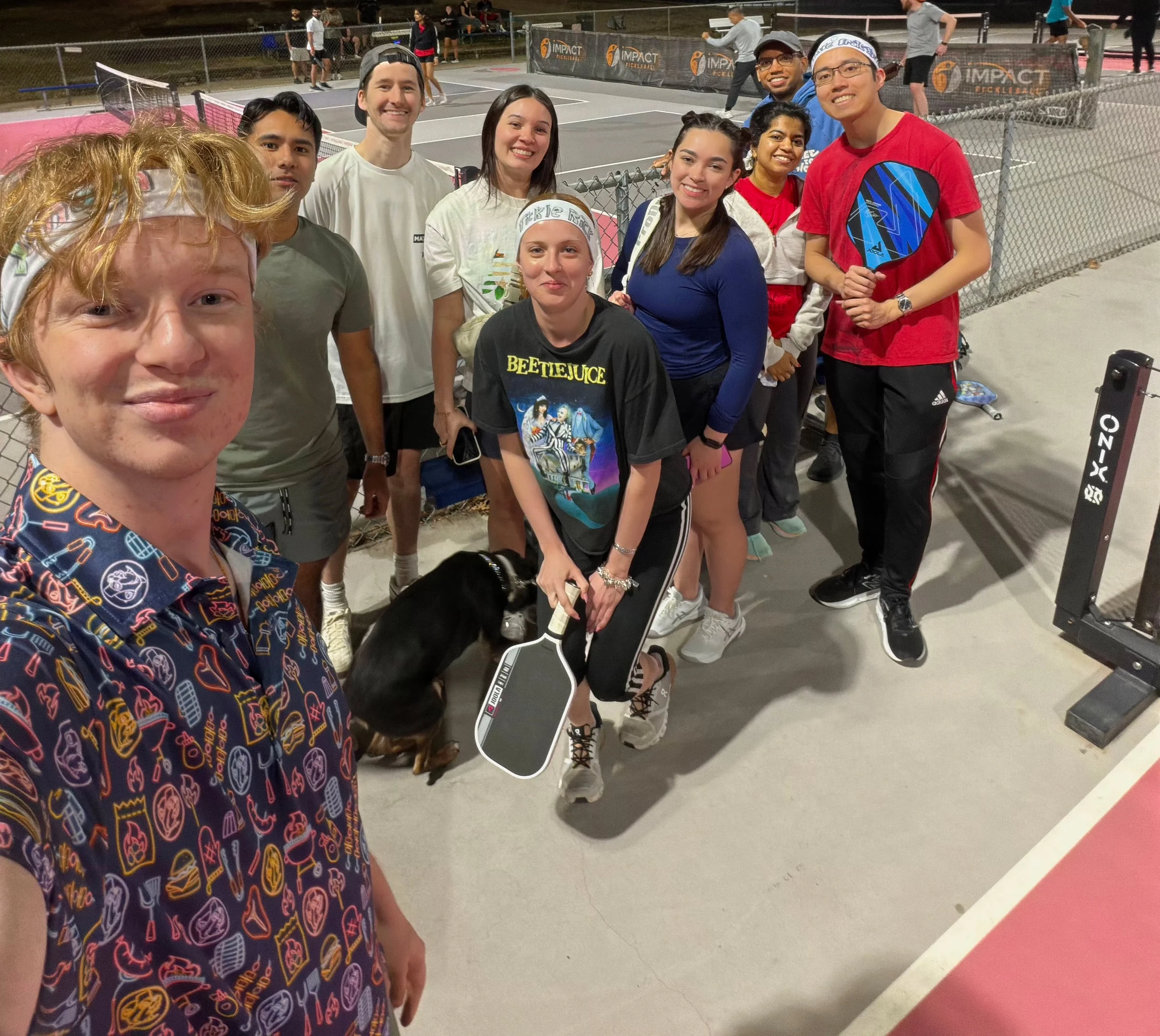 A group of friends and a black dog gathers for a selfie beside pickleball courts.
