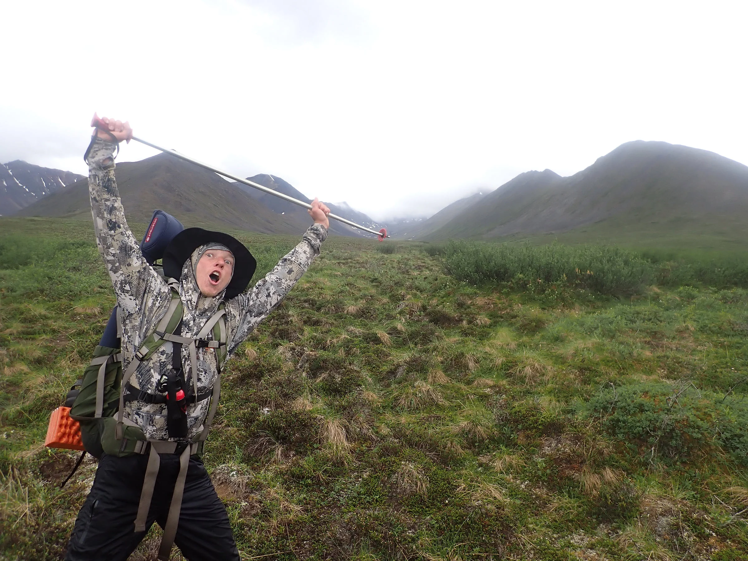 A backpacker throws a trekking pole overhead in a green mountain valley.