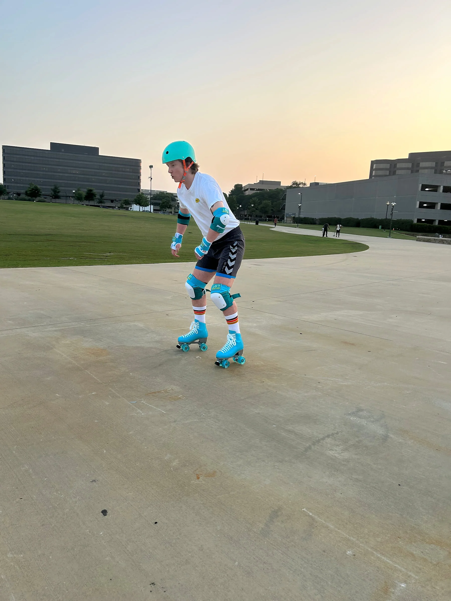 A roller skater in bright teal safety gear glides across a broad plaza at sunset.