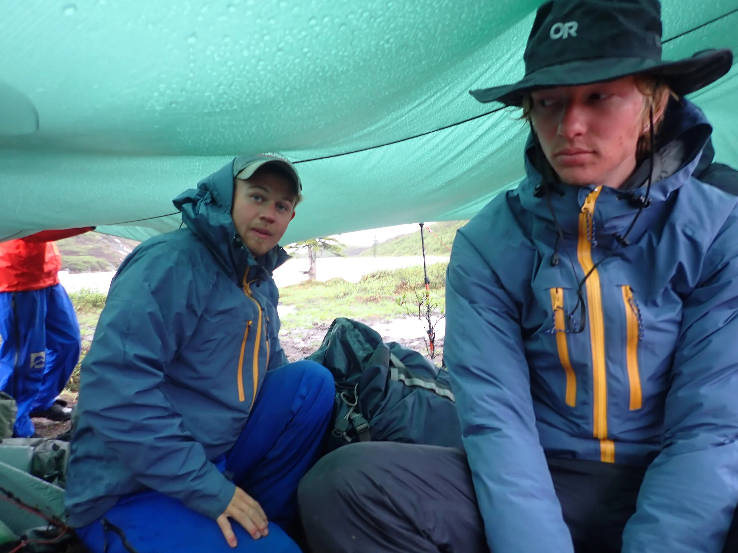 Two rain-soaked hikers sit beneath a teal tarp with their packs between them.